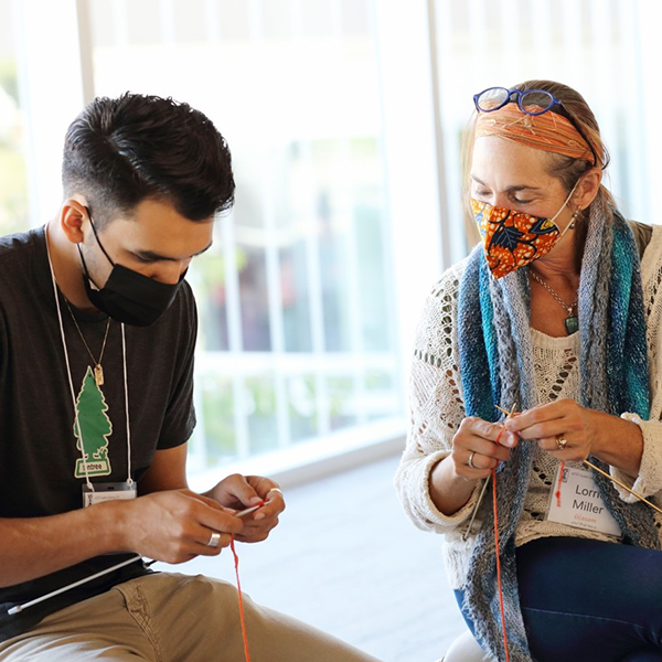 Dr. Lorrie Miller (right) helps a male student knit a tiny orange sweater at Indigenous Teachers Education Program – NITEP Day, September 24, 2021